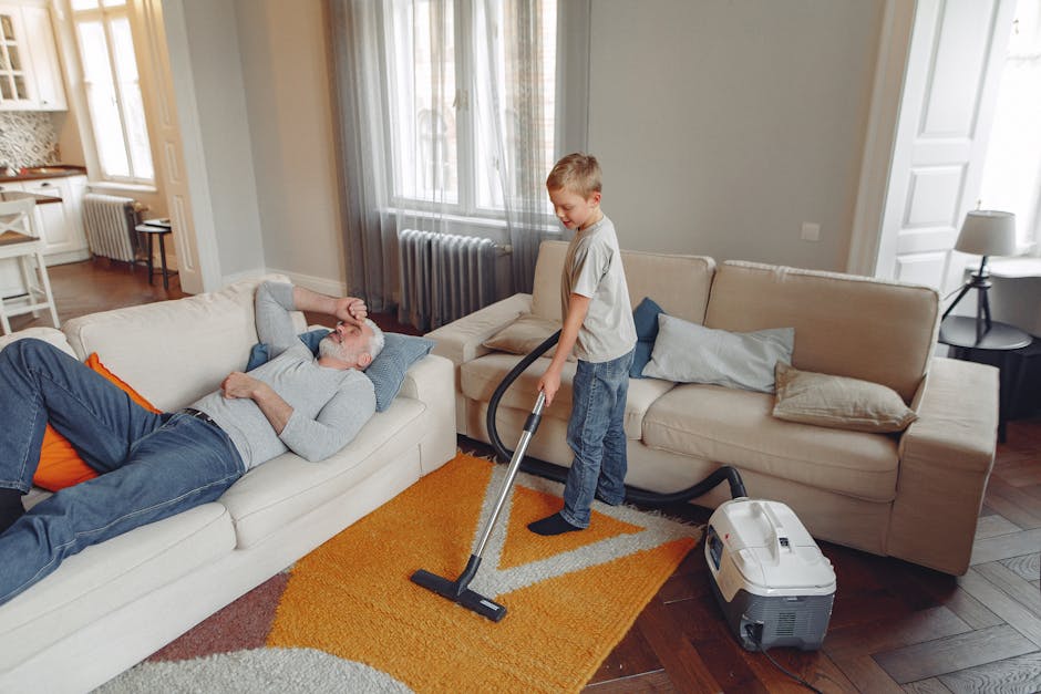 A young boy using a vacuum cleaner to clean the area rug in a living room with light-colored sofas, decorative pillows, and wooden flooring. An older man is lying on the sofa, appear to be resting with his hand on his forehead. The room features a large window with sheer curtains, a side table with a lamp, and a radiator against the wall. The scene illustrates domestic cleaning and surface maintenance, emphasizing the importance of deep cleaning and hygiene in a residential setting, with visible cleaning equipment from Earls Court Carpet Cleaners situated nearby.