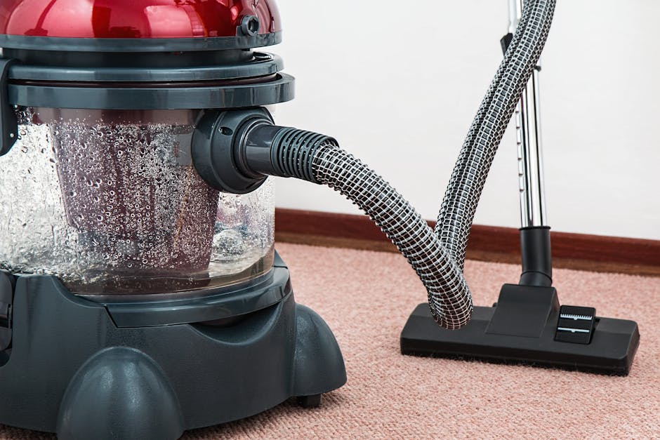 Close-up of a vacuum cleaner with a transparent dust container filled with dirt and debris, positioned on a pink carpeted floor in a residential or commercial indoor setting. The vacuum features a black base with a large, suction hose extending from the main unit, which is connected to a flexible, patterned hose. Bright lighting highlights the cleanliness of the environment, with a plain white wall and a wooden skirting board in the background. The scene emphasizes surface cleaning and maintenance, illustrating professional deep cleaning processes as offered by Earls Court Carpet Cleaners in Gloucester Road.