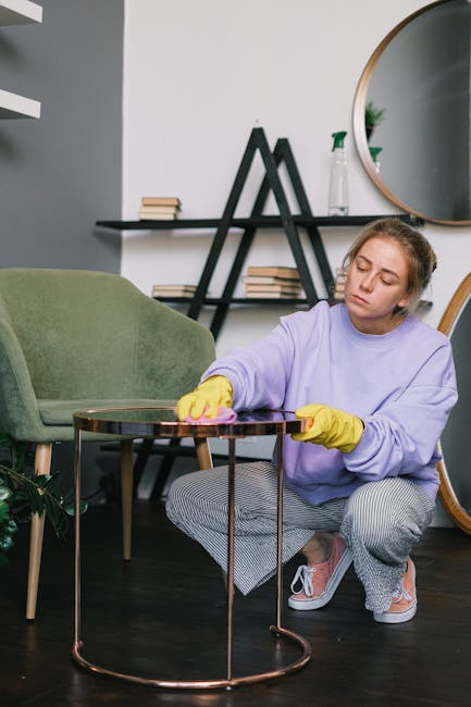 A person wearing gloves is using a handheld electric polishers on a dark gray fabric sofa in a living room. The sofa has a modern design with a high backrest and armrests, situated on a wooden floor. 
